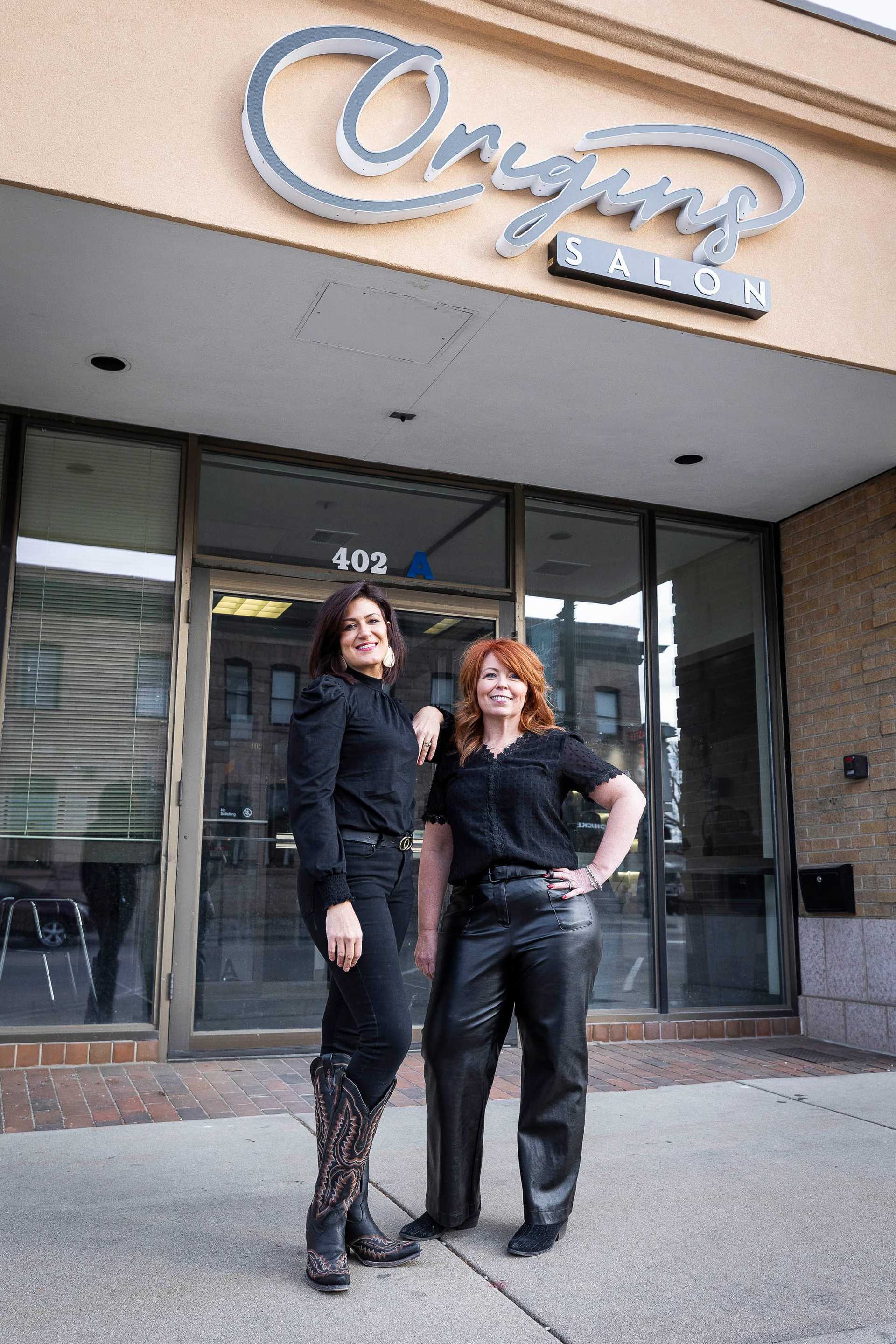 Two women in front of Origins Salon, smiling, wearing black outfits and boots.
