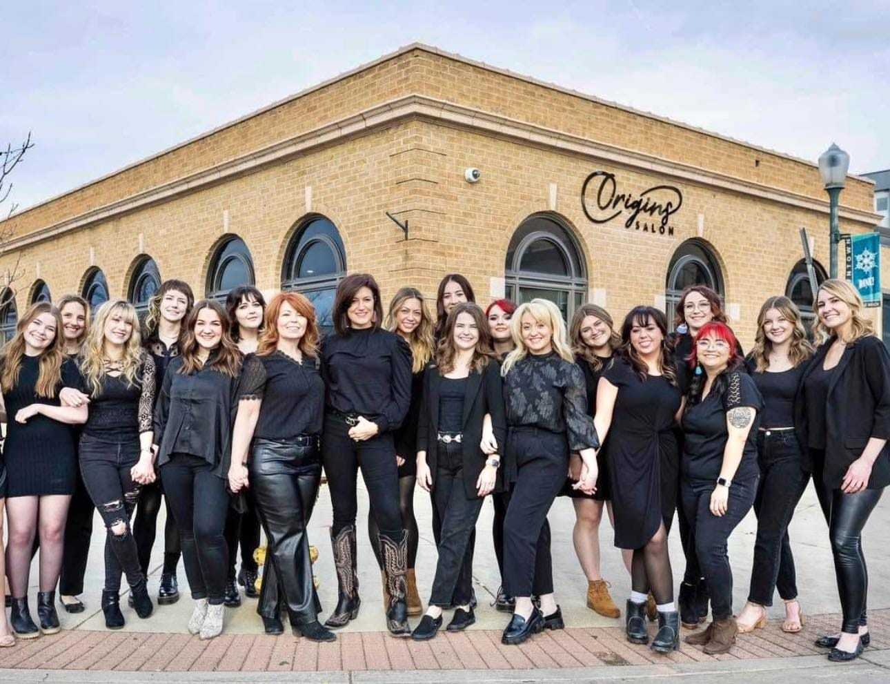 Team in black outfits posing outside Origins Salon building.