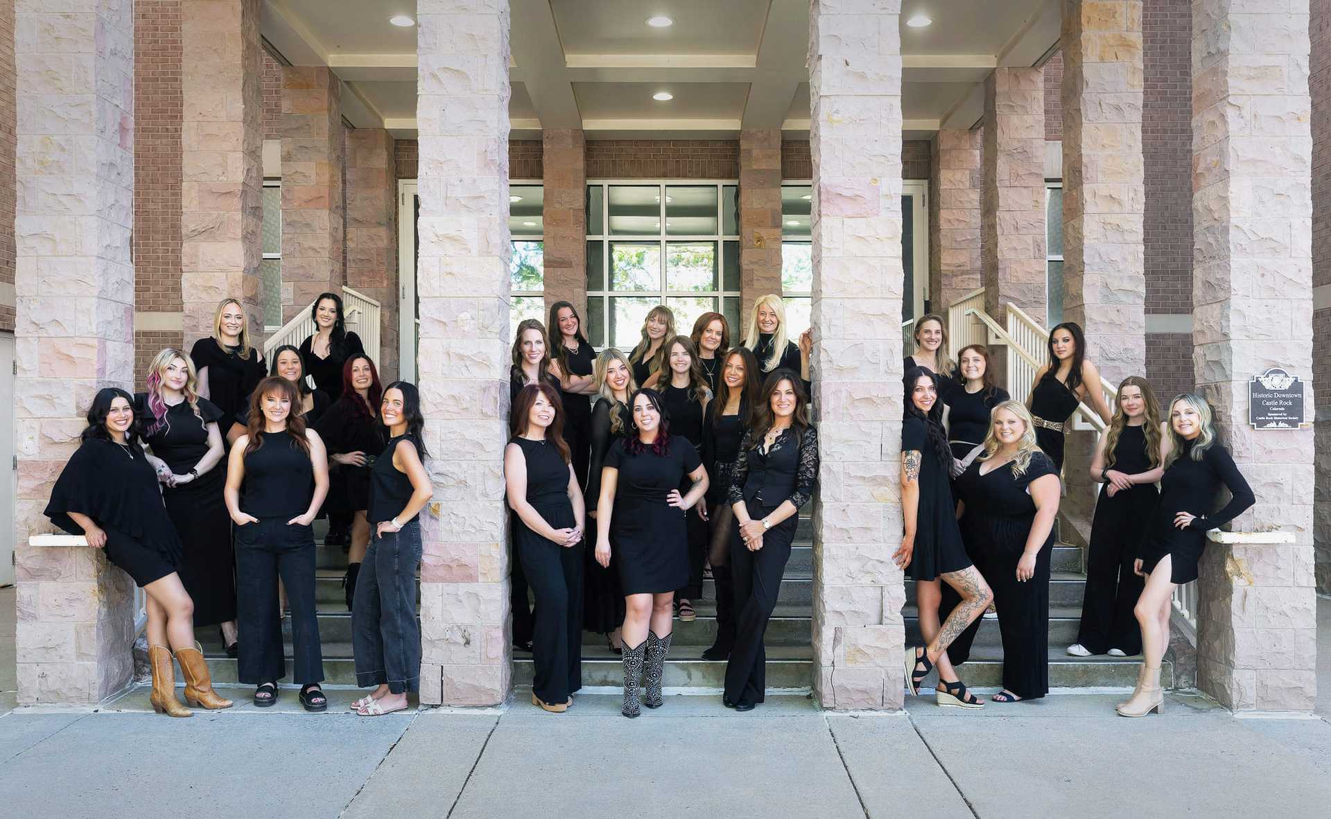 Group of women in black outfits posing on steps with stone pillars.