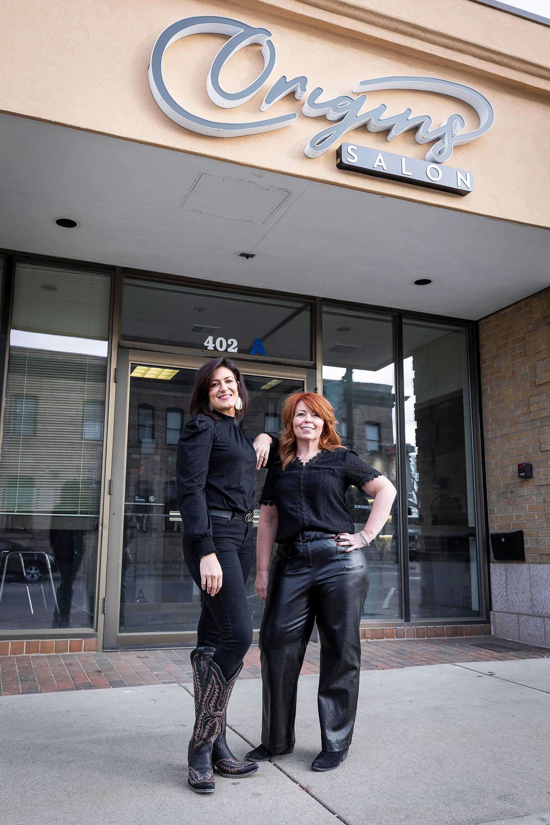 Two women in front of Origins Salon, smiling, wearing black outfits and boots.