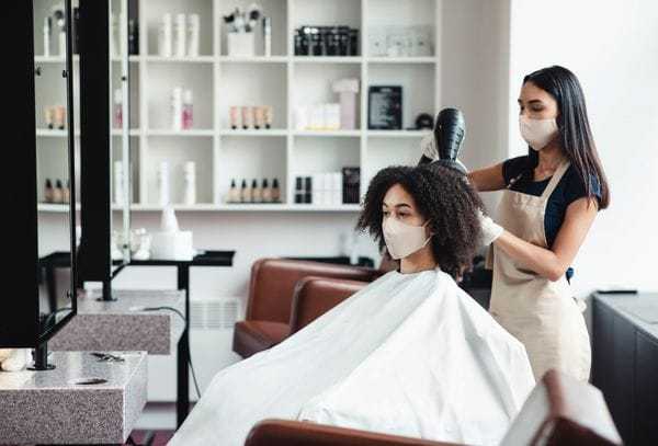 Hairstylist wearing a mask blow-drying a client's curly hair in a modern salon.